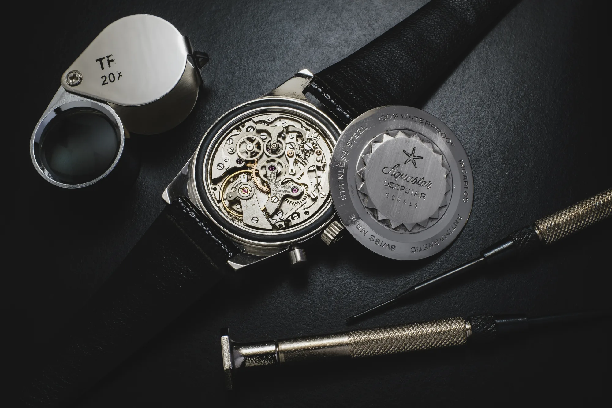 Vintage Aquastar Deepstar Chronograph mit abgenommenem Gehäuseboden, mit Blick auf das originale Säulenrad-Werk der 1960er-Jahre, daneben eine Uhrmacherlupe (20-fach), Schraubendreher und der gravierte Stahlgehäuseboden mit Prägung 100% Waterproof, Antimagnetic
