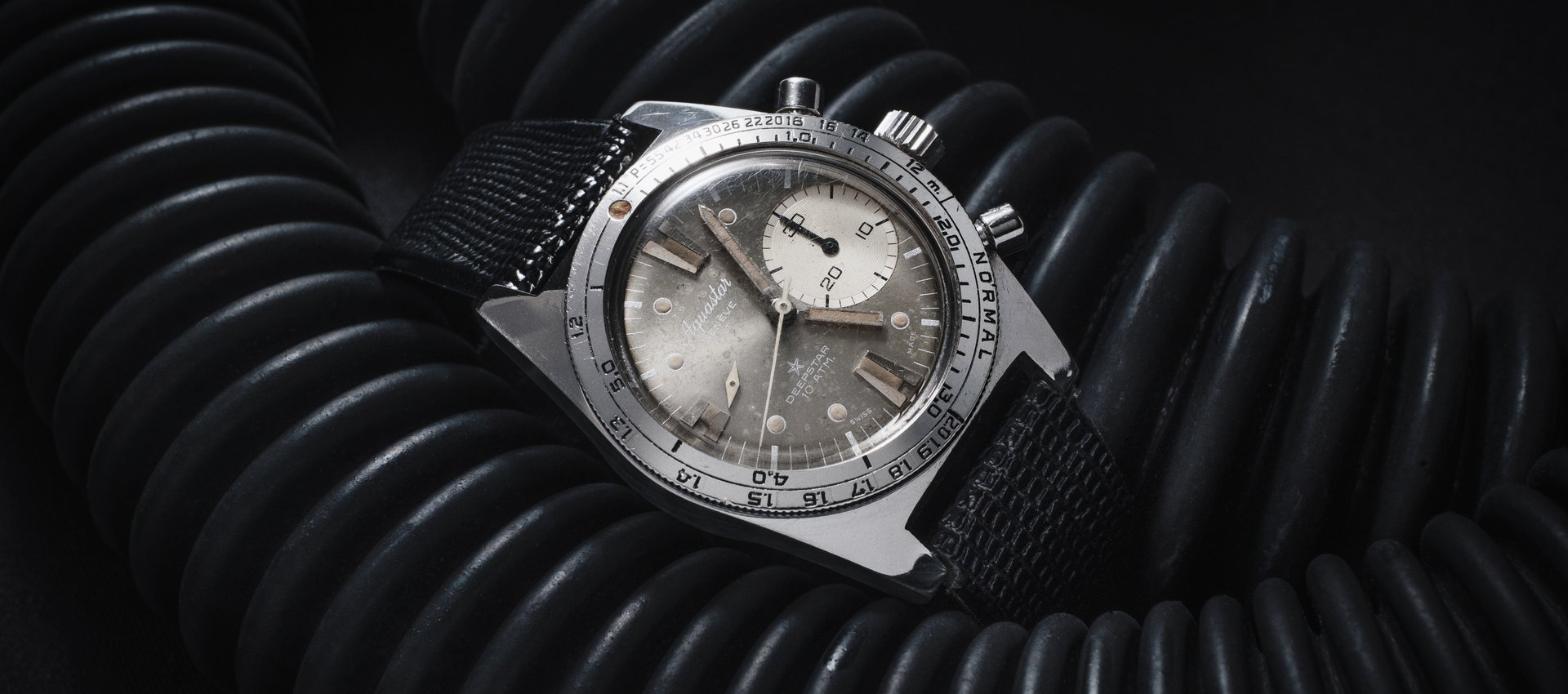 A vintage Aquastar Deepstar 10 ATM chronograph, silver dial with cream-aged tritium markers, resting on the black corrugated hoses of a period diving regulator