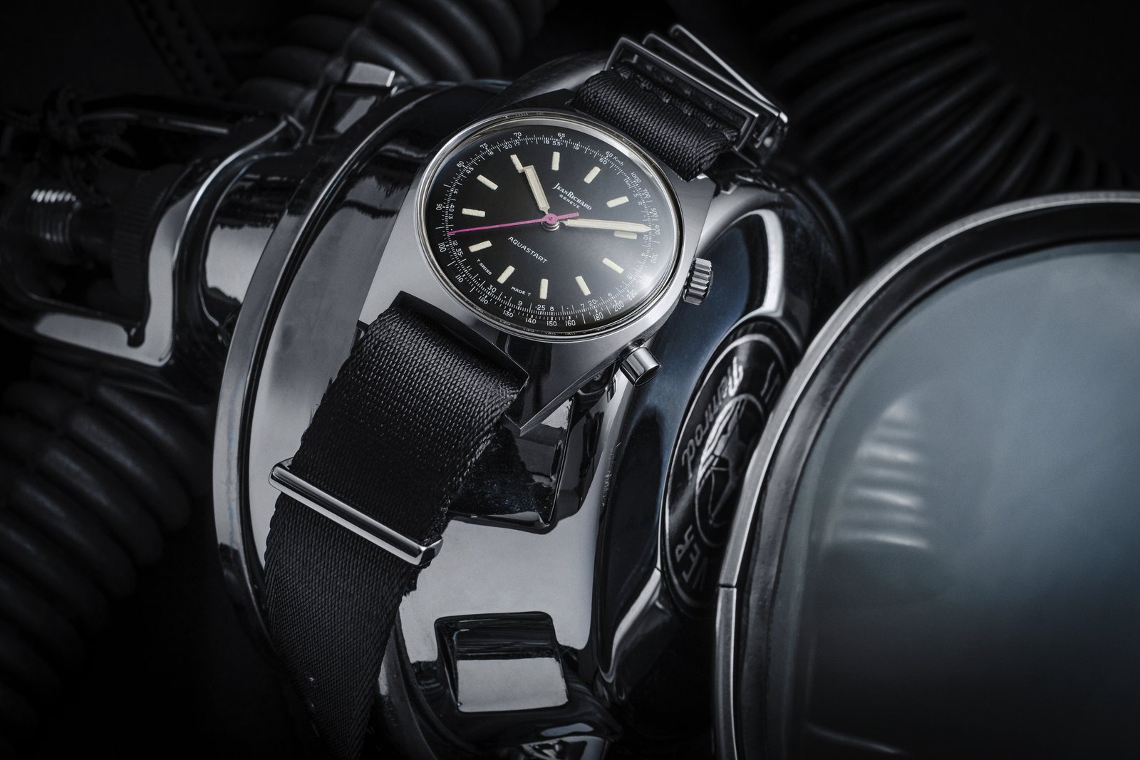 Vintage JeanRichard Geneve Aquastart yacht-timer wristwatch, black dial with kilometres-per-hour scale and magenta seconds hand, on black seatbelt-weave NATO strap next to chromed diving equipment